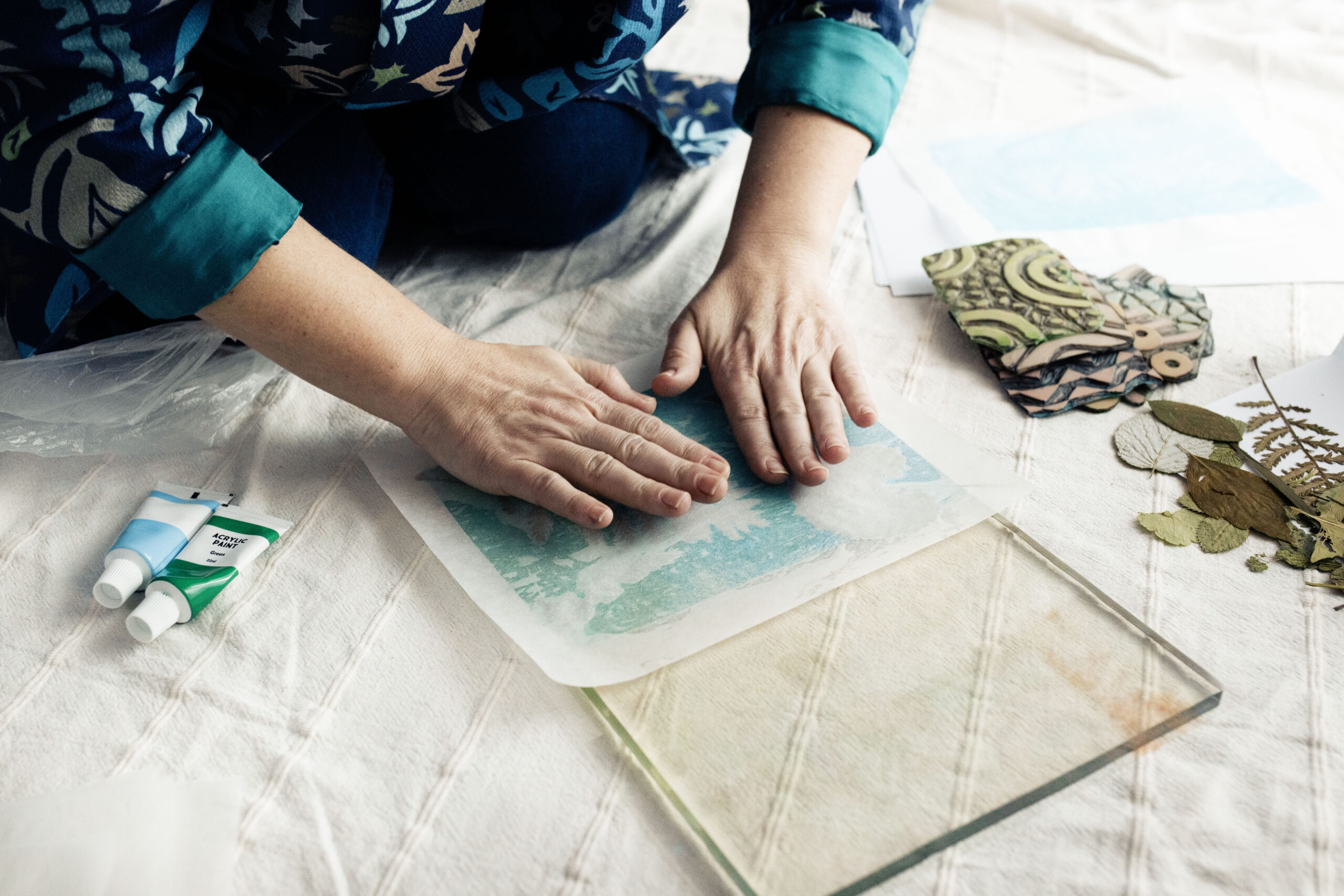 Hands of the founder and designer of Inner Imprints Arts Studio pressing a turquoise botanical print on a gelli printing plate using deli paper.