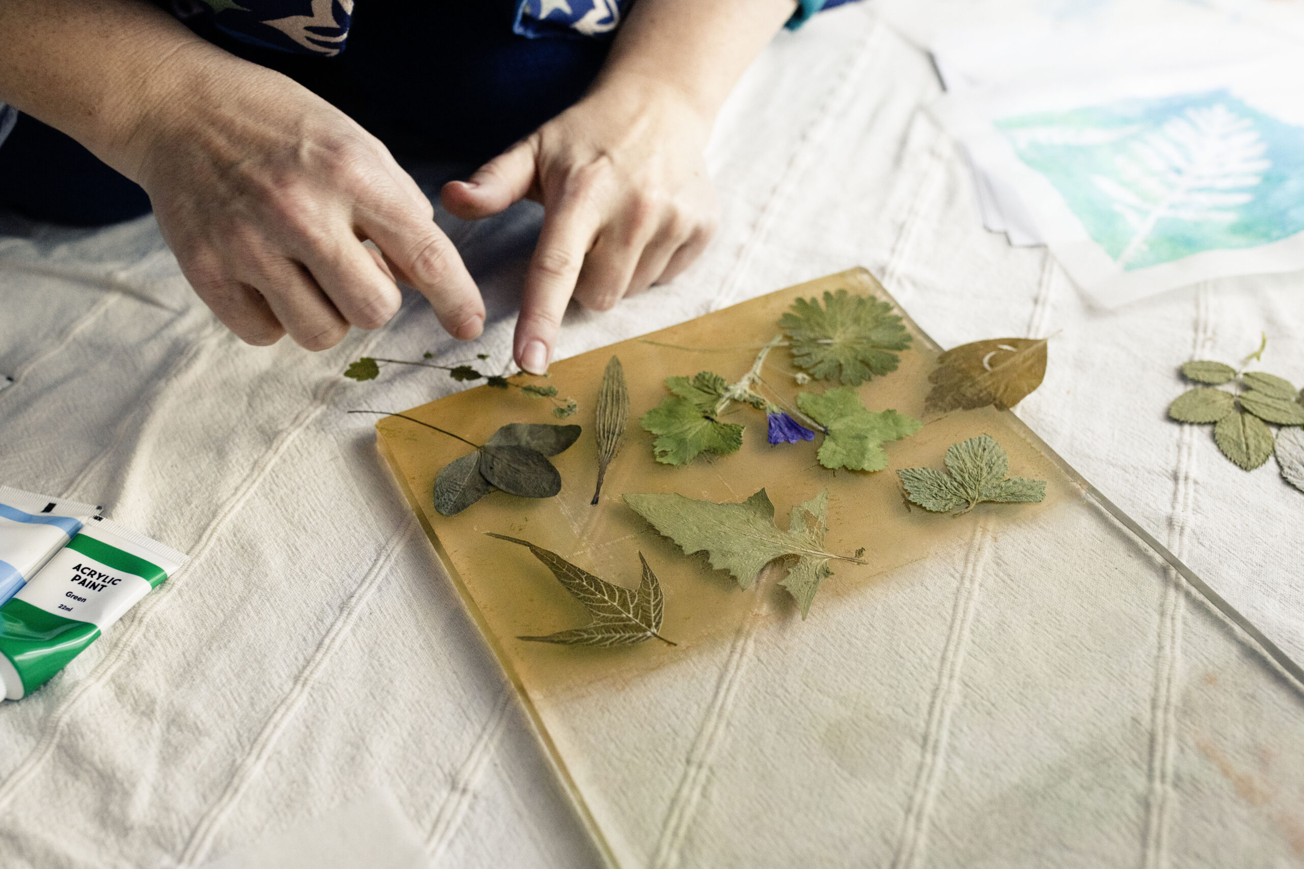Hands of the founder and designer of Inner Imprints Arts Studio arranging dried leaves and flowers on a transparent gelli printing plate.