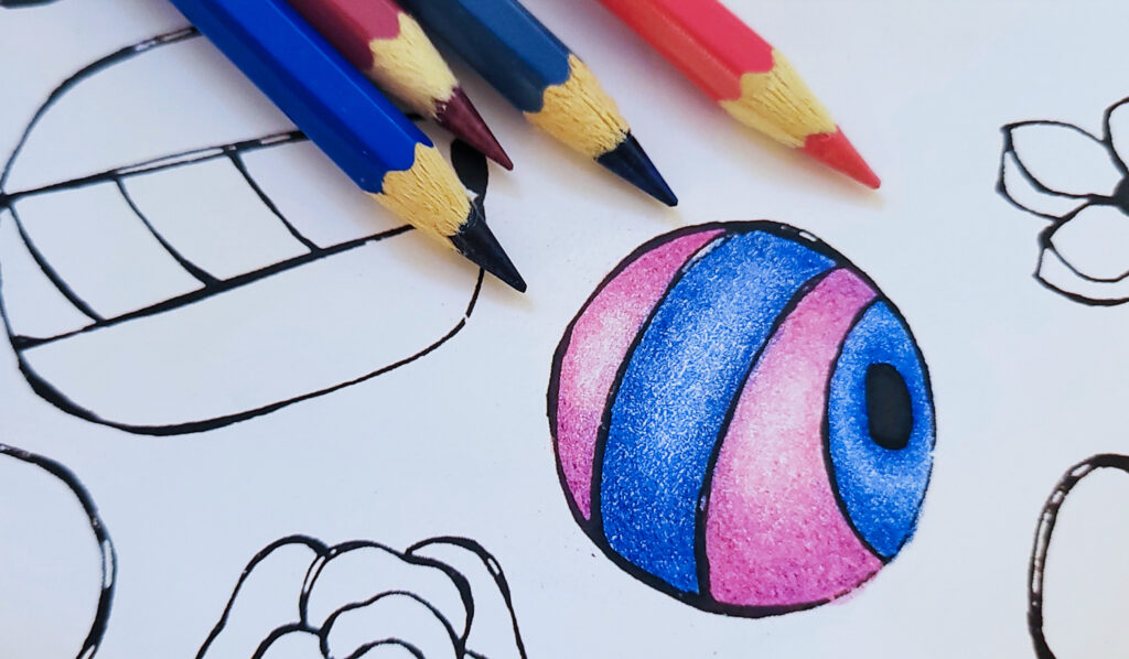 A close-up of a hand-coloured drawing showing a round bead shaded in pink and blue with coloured pencils lying nearby, colouring by Inner Imprints.