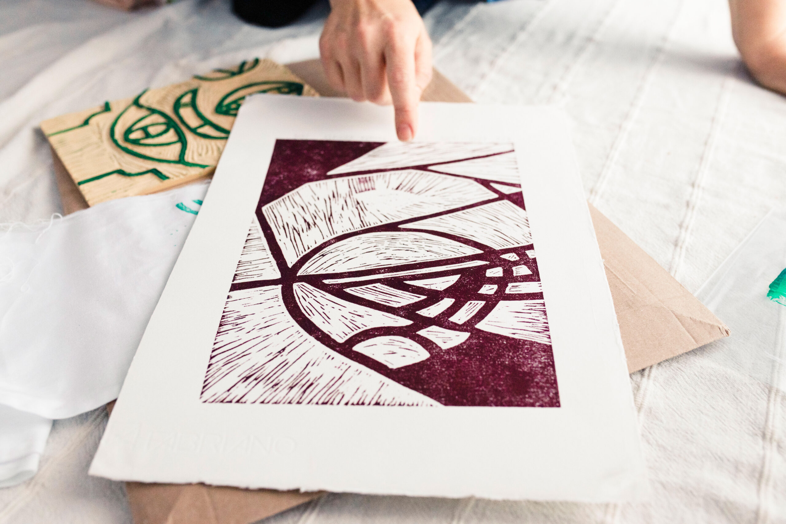 Renata, founder and designer of Inner Imprints, points to her burgundy linocut print lying on a white linen background. The carved linoleum block is visible beside it, showing the process behind block printing.