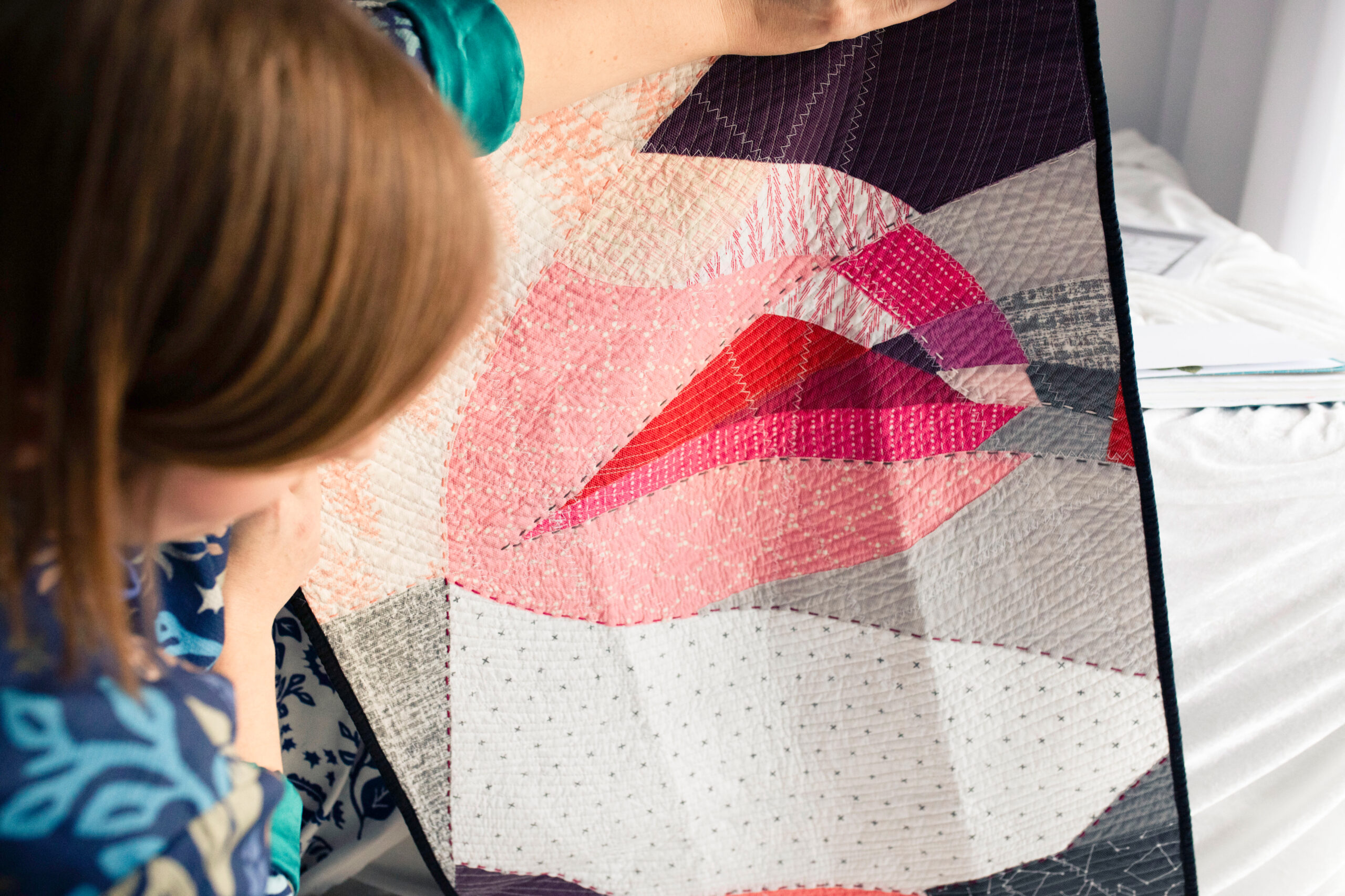 Renata, the founder and designer of Inner Imprints, holds up her own quilted wall hanging made of geometric fabric pieces in shades of pink, magenta, orange-red, peach, grey, and white. The quilt features an abstract representation of lips. She examines the handmade piece closely while standing next to a window that brightens up the room.